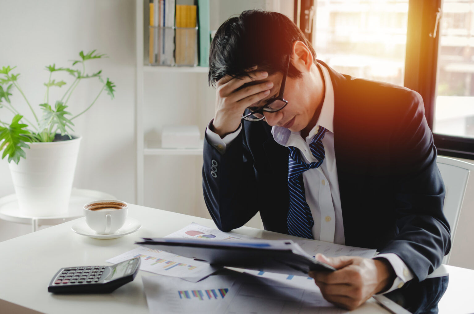 There is an headache and tired stressed asian young employee businessman working finance about cost with clipboard document report and coffee cup on desk at home office, illustrating customer relationship management software