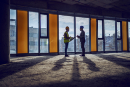 ORM and outsourcing relationship management software illustration: silhouette of construction worker and architect standing near window in future business center and talking about realization of the project.